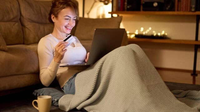 A smiling woman sits on the floor covered with a blanket, holding a credit card and using a laptop. A mug is on the floor nearby, and warm lights decorate the background, creating a cozy atmosphere.