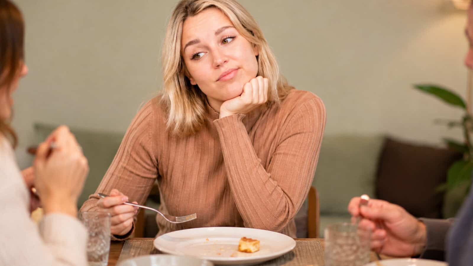 A woman with blonde hair rests her chin on her hand and looks bored or unimpressed while sitting at a table with an empty plate, surrounded by two other people eating.