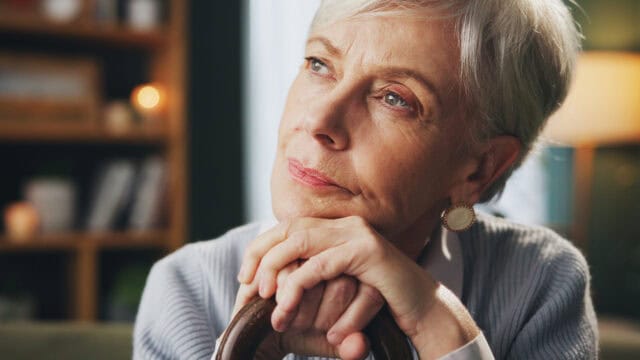 Elderly woman with short gray hair gazes thoughtfully into the distance, resting her chin on her hands while sitting indoors. Warm lighting and bookshelves appear blurred in the background.
