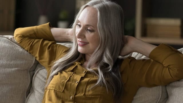 A smiling older woman with long gray hair sits on a beige couch, wearing a mustard yellow shirt and resting her arms behind her head, appearing relaxed and content. Shelves with books are blurred in the background.