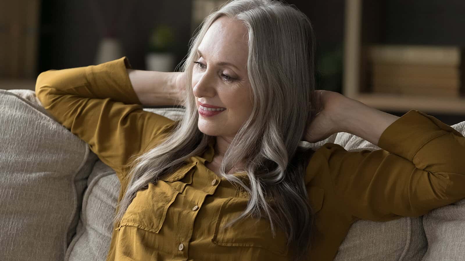 A smiling older woman with long gray hair sits on a beige couch, wearing a mustard yellow shirt and resting her arms behind her head, appearing relaxed and content. Shelves with books are blurred in the background.