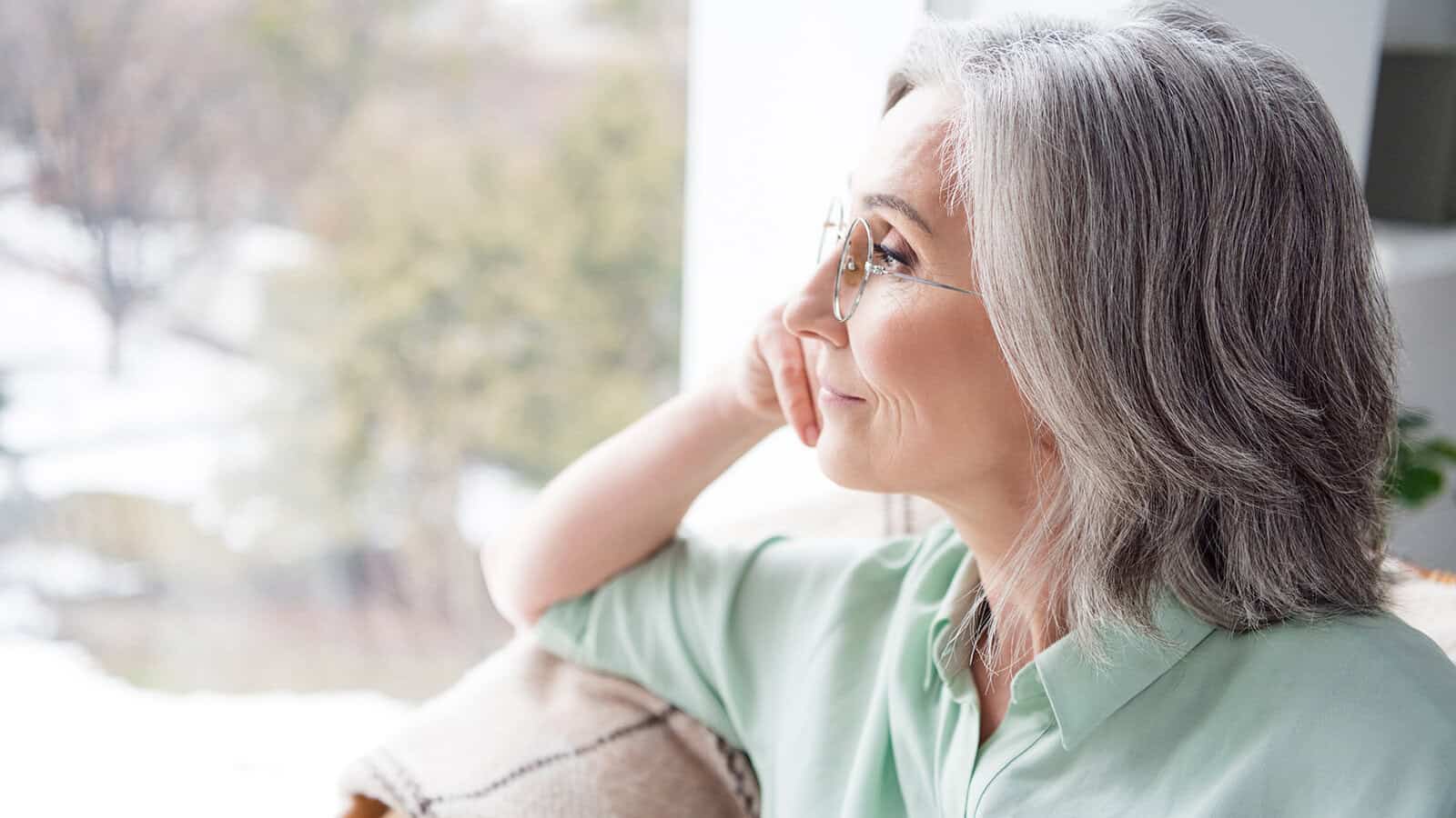 Older woman with gray hair and glasses sits indoors by a window, looking outside thoughtfully. She is wearing a light green shirt and resting her chin on her hand. Snow and trees are visible outside.