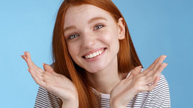 A smiling young woman with long red hair and freckles, wearing a striped shirt, holds her hands up near her face against a blue background.