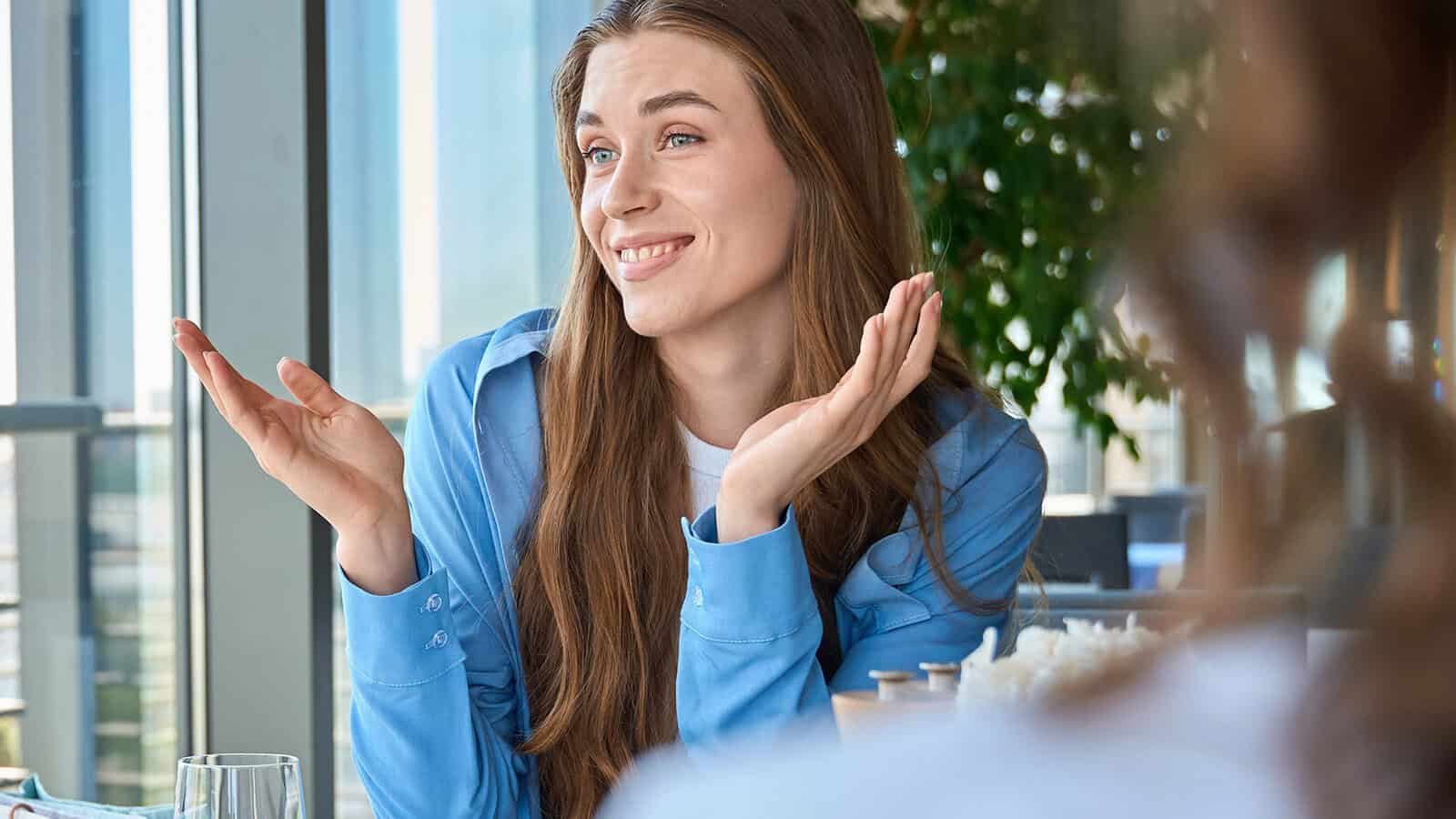 A woman with long brown hair, wearing a blue shirt, smiles and shrugs with both hands raised, sitting indoors near a window with greenery in the background.