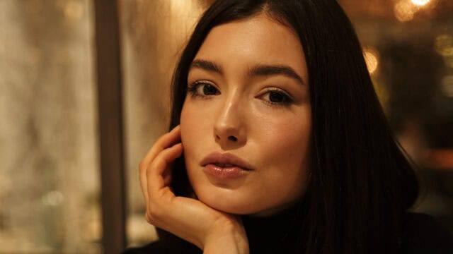 A person with long dark hair rests their chin on their hand, looking directly at the camera with a calm expression. The background is softly blurred with warm lighting.