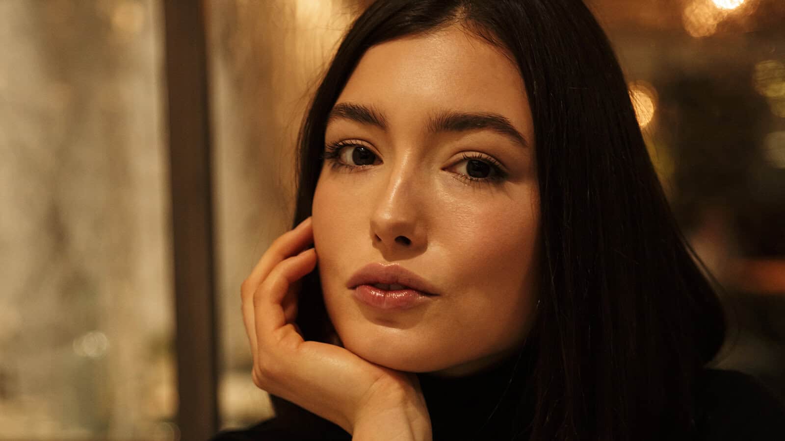 A person with long dark hair rests their chin on their hand, looking directly at the camera with a calm expression. The background is softly blurred with warm lighting.