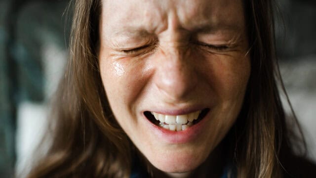 Close-up of a woman with long brown hair, eyes shut tightly, and mouth open, appearing to be in pain or distress.