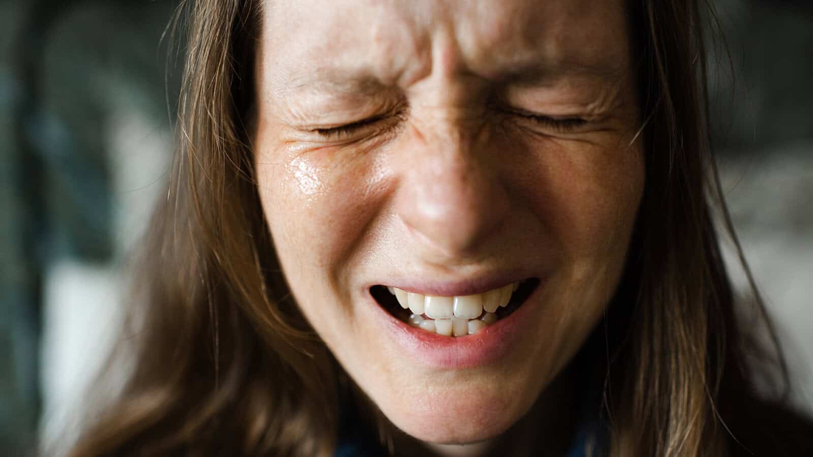 Close-up of a woman with long brown hair, eyes shut tightly, and mouth open, appearing to be in pain or distress.