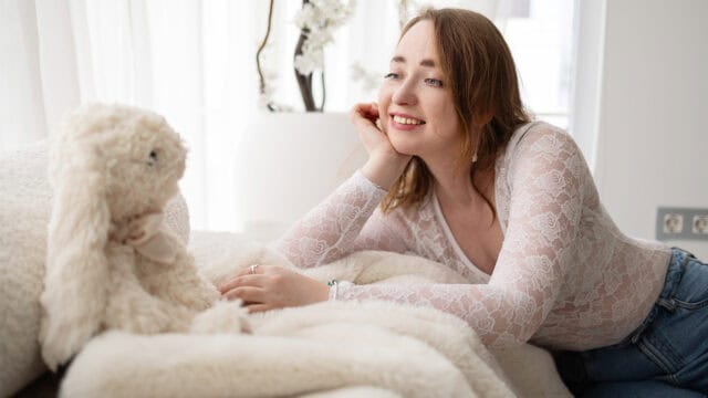 A woman wearing a white lace top smiles while lying on a bed, looking at a plush bunny toy. The room is bright with natural light and decorated in soft, neutral colors.