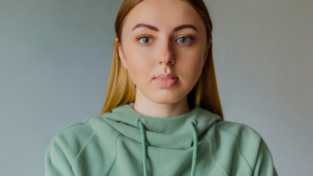 A young person with long straight blonde hair wearing a light green hoodie looks directly at the camera against a plain background, suggesting an introspective moment that hints at their unique personality traits.