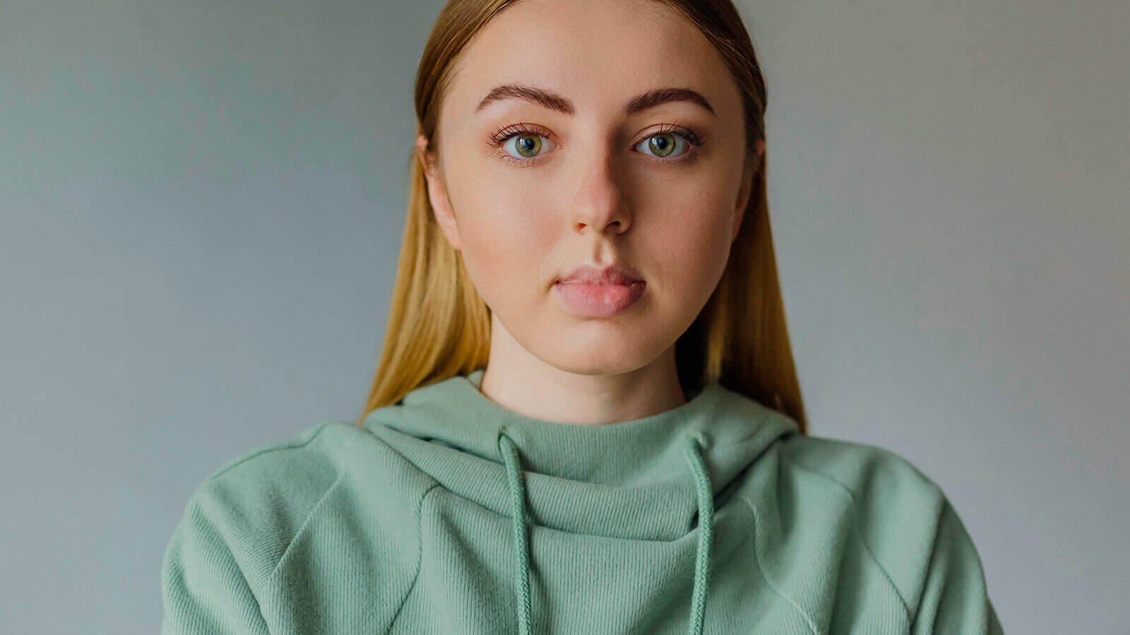 A young person with long straight blonde hair wearing a light green hoodie looks directly at the camera against a plain background, suggesting an introspective moment that hints at their unique personality traits.