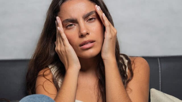 A woman with long brown hair sits on a couch, holding her face with both hands and looking ahead with a concerned or stressed expression.