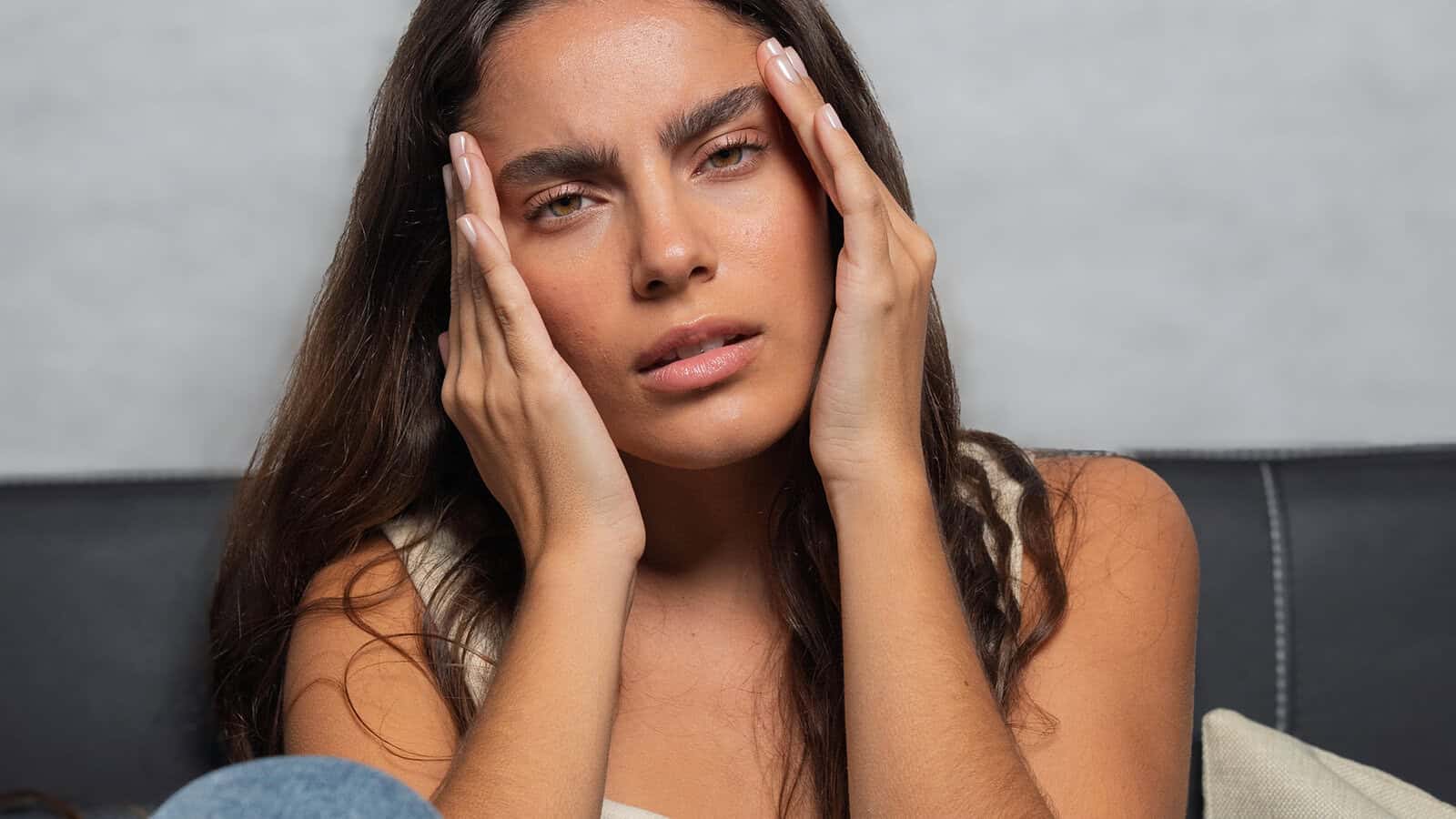 A woman with long brown hair sits on a couch, holding her face with both hands and looking ahead with a concerned or stressed expression.