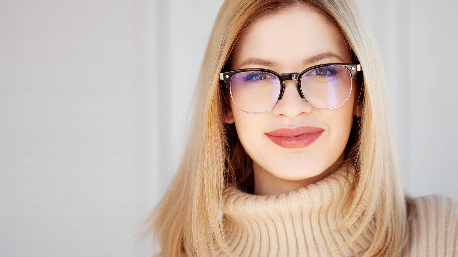 A woman with long blonde hair and black-rimmed glasses smiles at the camera. She is wearing a beige turtleneck sweater, and the background is softly blurred in light tones.