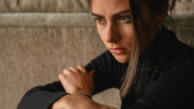 A woman with long dark hair sits with her arms crossed on her knees, looking thoughtful and serious. She wears a black ribbed sweater and gazes off to the side in soft lighting.