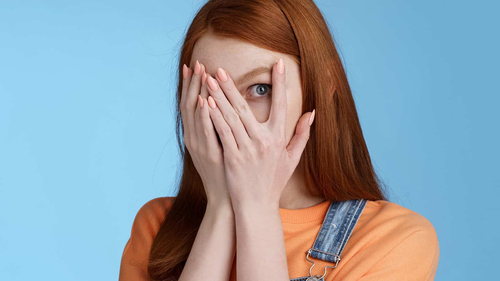 A person with long red hair covers most of their face with their hands, peeking through their fingers. They wear an orange top and denim overalls, with a light blue background.