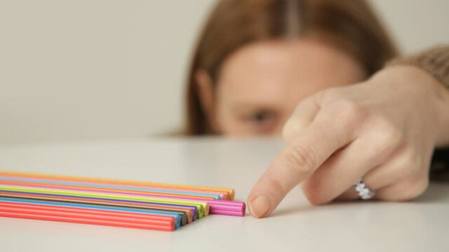 A person points at a pink straw slightly out of alignment with a neat row of colorful straws on a white surface.