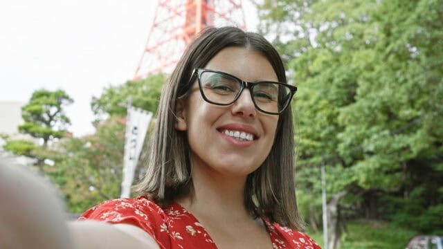 A woman with dark hair and glasses, wearing a red floral top, smiles at the camera outdoors with trees and a tall red structure in the background.