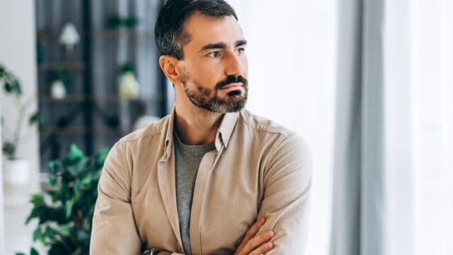 A man with short dark hair and a beard stands indoors with his arms crossed, looking to his left. He wears a beige shirt over a gray t-shirt. The background is softly lit with plants and curtains visible.