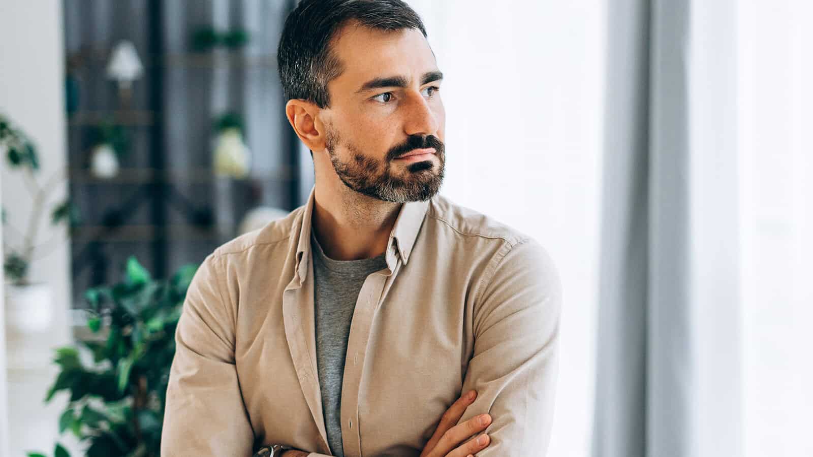 A man with short dark hair and a beard stands indoors with his arms crossed, looking to his left. He wears a beige shirt over a gray t-shirt. The background is softly lit with plants and curtains visible.