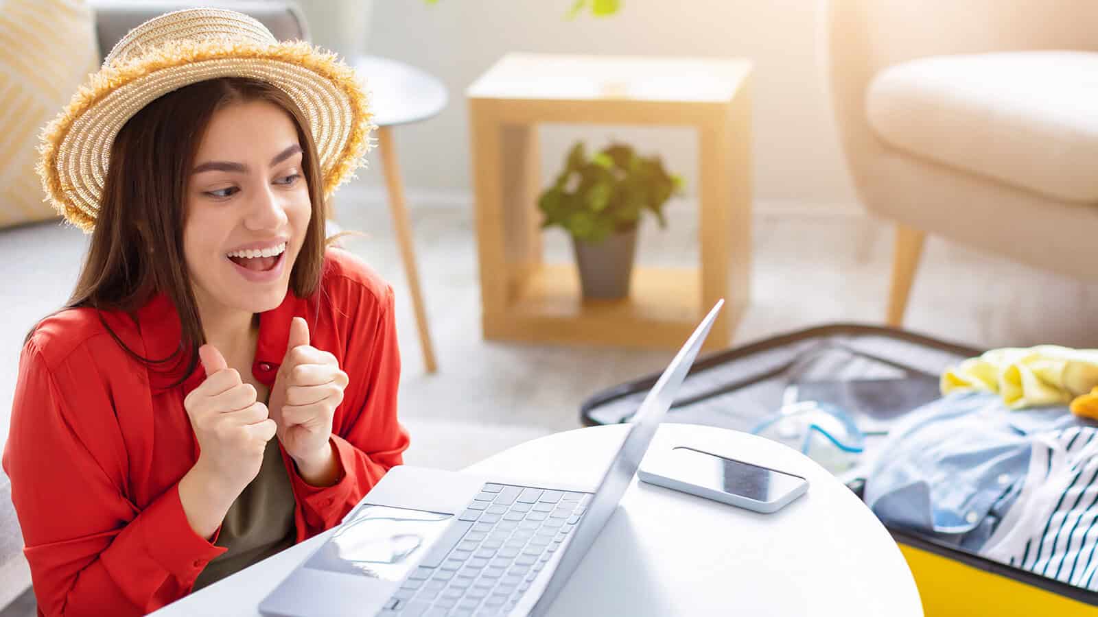 A smiling woman in a straw hat sits at a table with a laptop, giving two thumbs up. An open suitcase and a phone are nearby, suggesting she is preparing for travel. Sunlight brightens the cozy room.