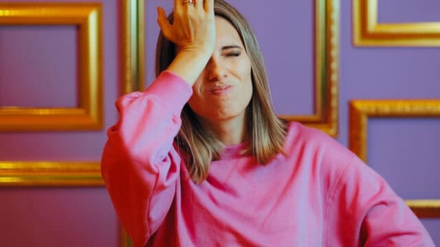 A woman in a pink sweatshirt makes a facepalm gesture, pressing her hand to her forehead and grimacing, standing in front of a purple wall decorated with gold-framed rectangles.