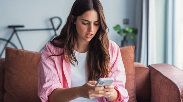 A young woman with long brown hair sits on a couch, looking surprised or concerned while reading something on her smartphone. A blurred bicycle and a plant are visible in the background.
