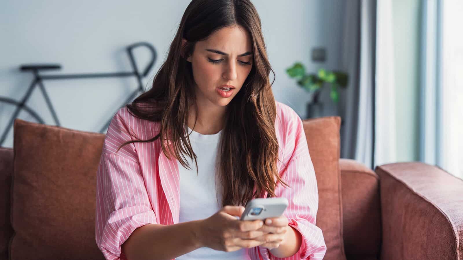 A young woman with long brown hair sits on a couch, looking surprised or concerned while reading something on her smartphone. A blurred bicycle and a plant are visible in the background.