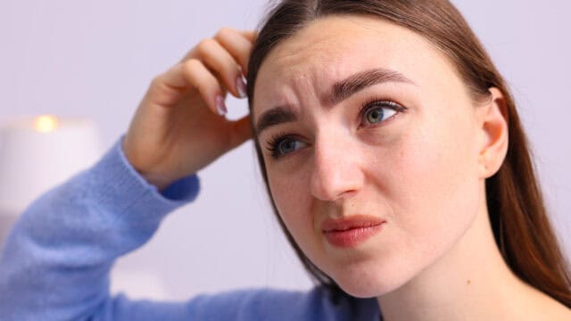A young woman with long brown hair and a blue sweater looks confused or thoughtful, scratching her head and furrowing her brow.
