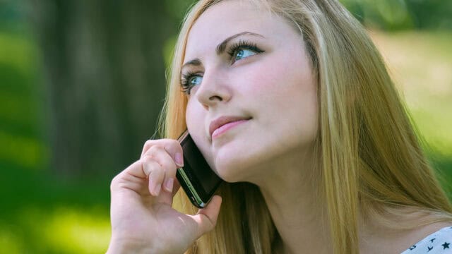 A woman with long blonde hair holds a smartphone to her ear, looking thoughtful as she talks on the phone outdoors in a green, blurred background.