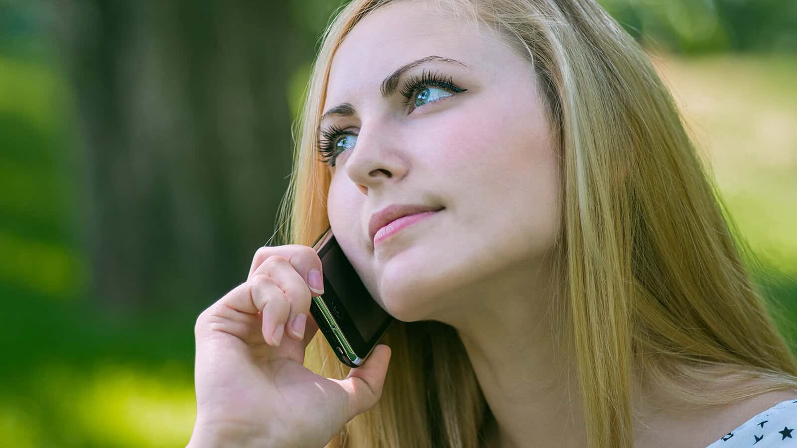 A woman with long blonde hair holds a smartphone to her ear, looking thoughtful as she talks on the phone outdoors in a green, blurred background.