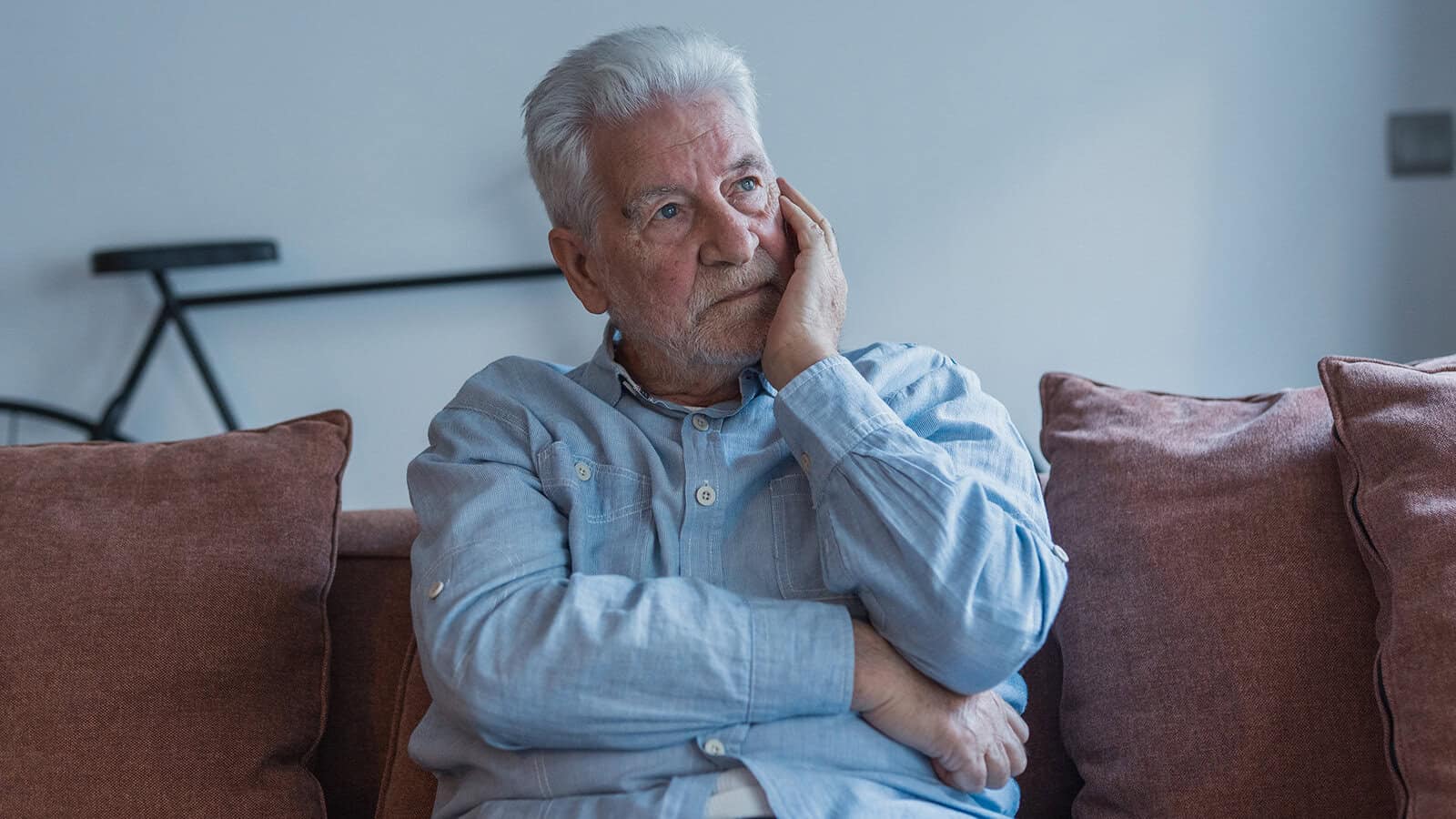 An elderly man with white hair sits on a couch with his arms crossed, resting his face on his hand and looking thoughtfully into the distance, perhaps contemplating spending in retirement and reflecting on the psychology of saving that shapes the lives of many retirees in their 70s.