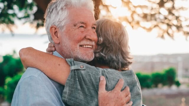 An older man smiles while hugging a woman outdoors at sunset. They both have gray hair and appear happy, with greenery and soft sunlight in the background.