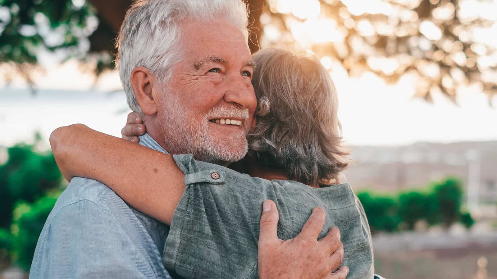 An older man smiles while hugging a woman outdoors at sunset. They both have gray hair and appear happy, with greenery and soft sunlight in the background.