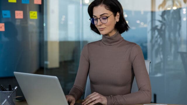 A woman with short dark hair and glasses works on a laptop at a desk in a modern office with glass walls and colorful sticky notes visible in the background.