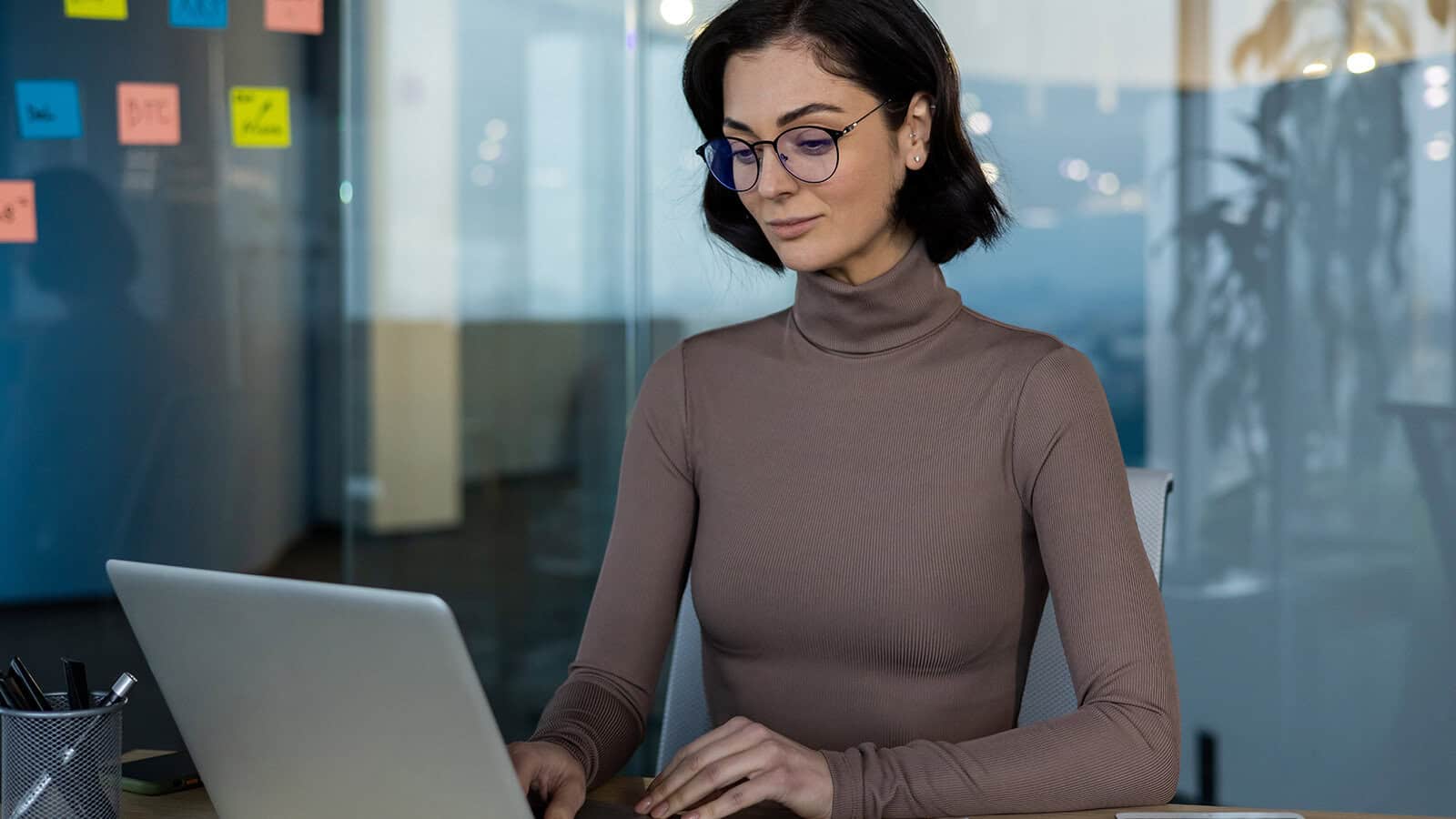 A woman with short dark hair and glasses works on a laptop at a desk in a modern office with glass walls and colorful sticky notes visible in the background.