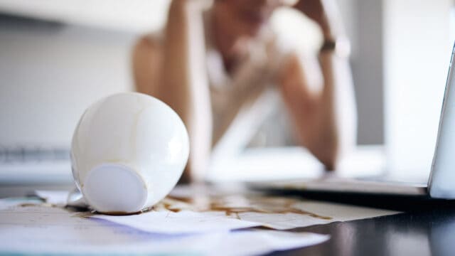A spilled coffee cup lies on papers, with coffee soaking into them. In the blurred background, a person sits at a desk with their head in their hands, looking stressed.