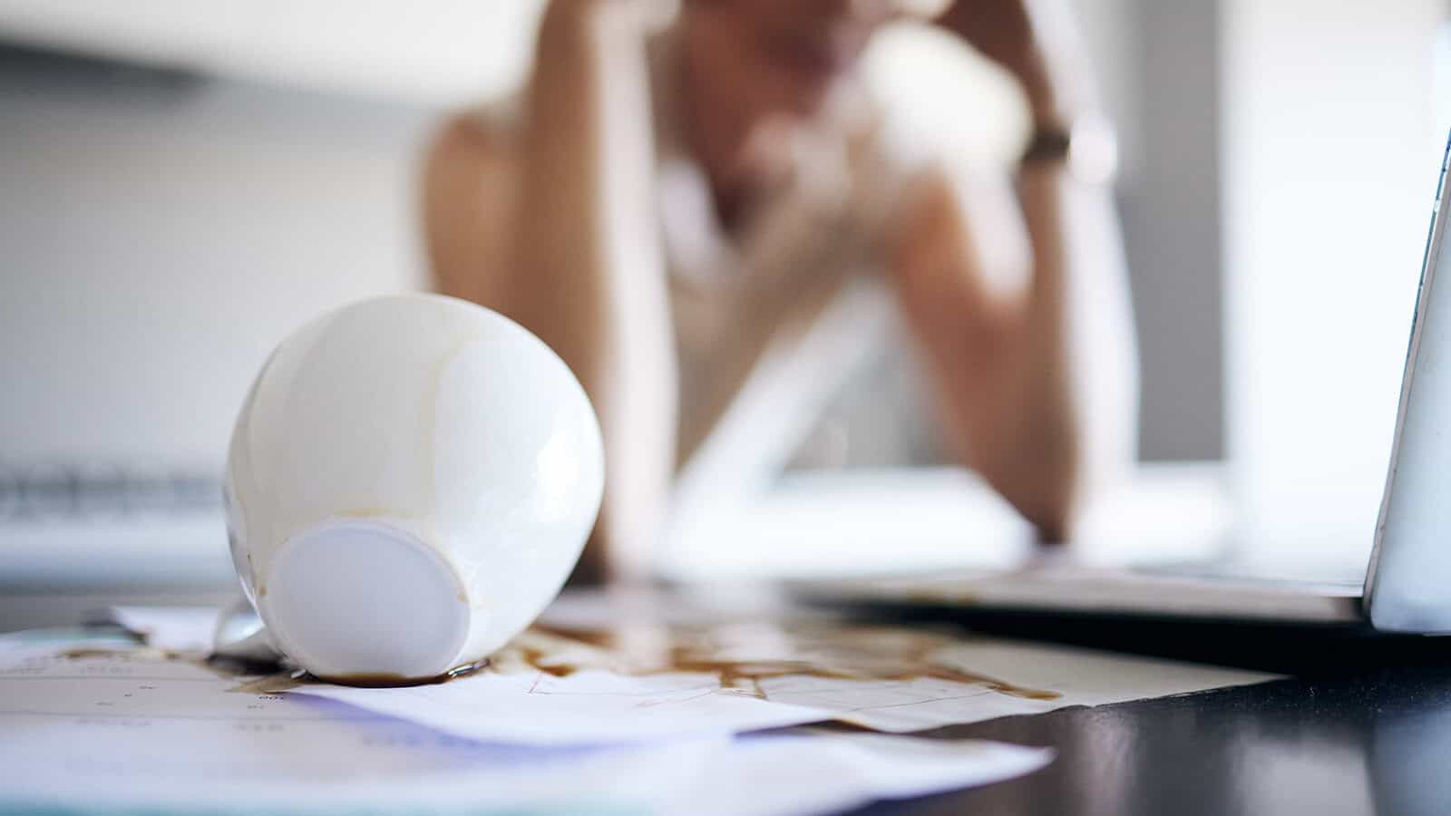 A spilled coffee cup lies on papers, with coffee soaking into them. In the blurred background, a person sits at a desk with their head in their hands, looking stressed.