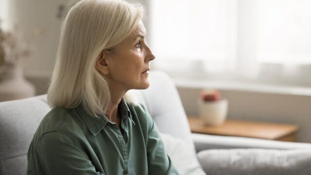 An older woman with gray hair sits on a couch, looking thoughtfully to the side in a softly lit room.