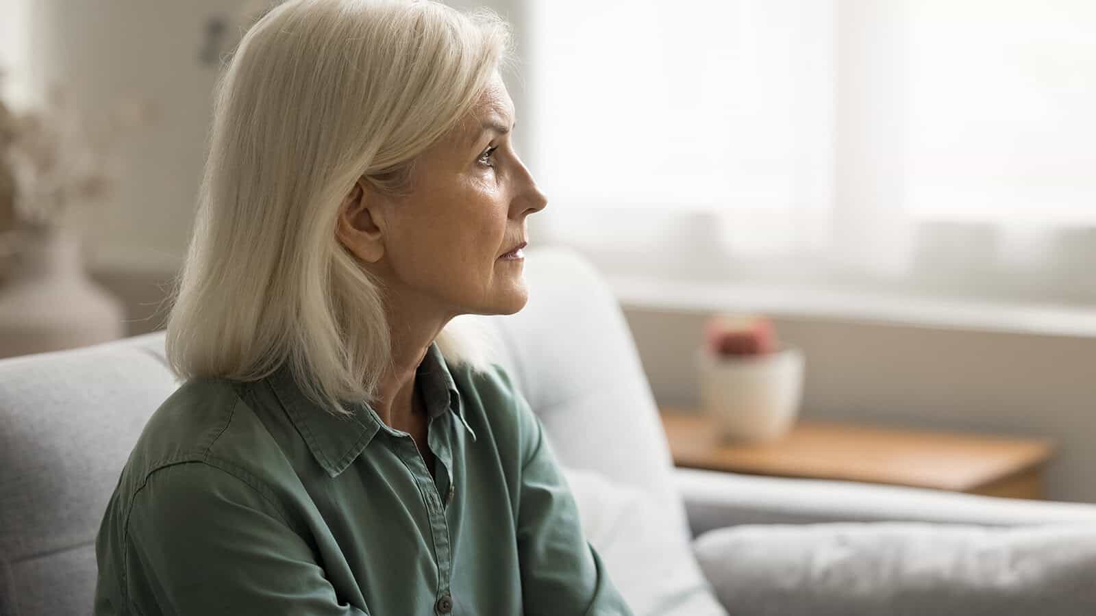 An older woman with gray hair sits on a couch, looking thoughtfully to the side in a softly lit room.