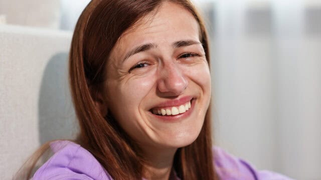 A young woman with long brown hair smiles while tears run down her cheeks, suggesting she is crying from laughter or experiencing mixed emotions. She is wearing a purple shirt and sitting indoors.