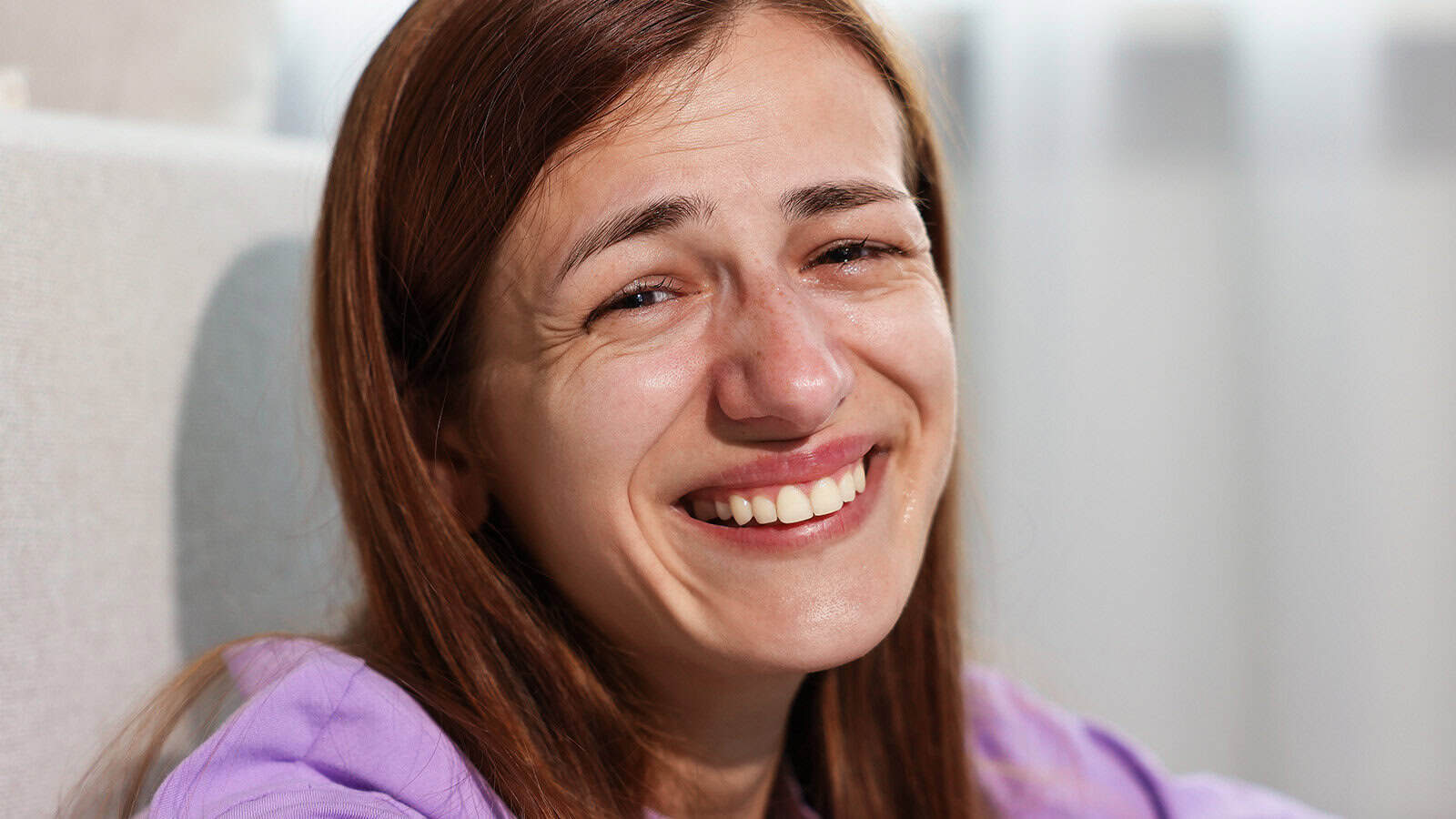 A young woman with long brown hair smiles while tears run down her cheeks, suggesting she is crying from laughter or experiencing mixed emotions. She is wearing a purple shirt and sitting indoors.