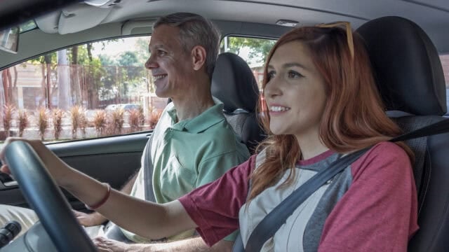 A young woman drives a car with an older man in the passenger seat. Both are smiling, and the car is parked outdoors on a sunny day.