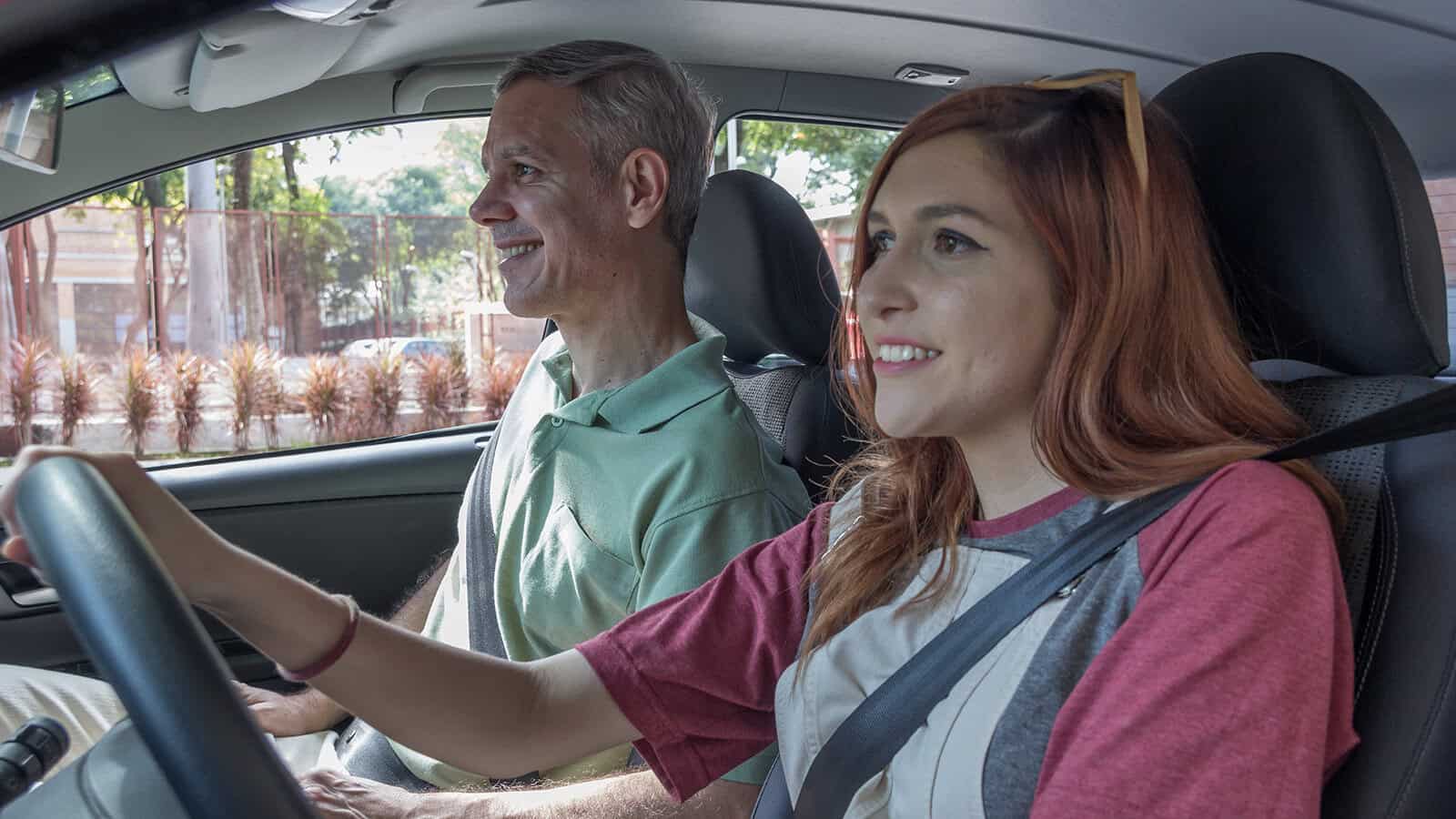 A young woman drives a car with an older man in the passenger seat. Both are smiling, and the car is parked outdoors on a sunny day.