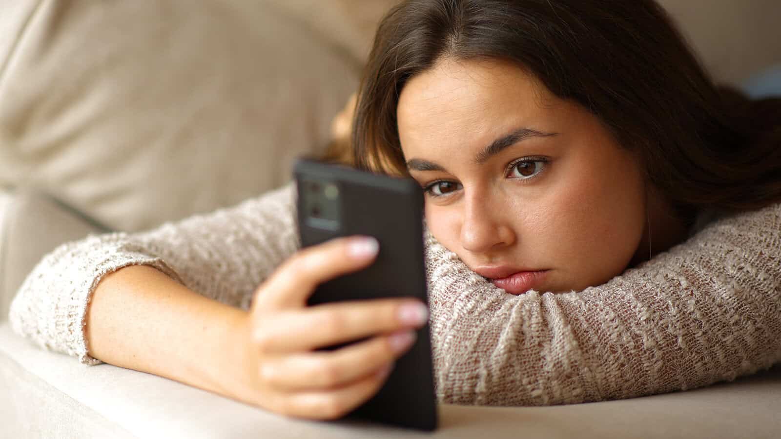 A young woman lies on a couch, resting her head on her arm, and looks intently at her smartphone with a serious, thoughtful expression—a quiet moment that reflects the modern life exhaustion brought on by constant connectivity and the never-ending demands for attention.