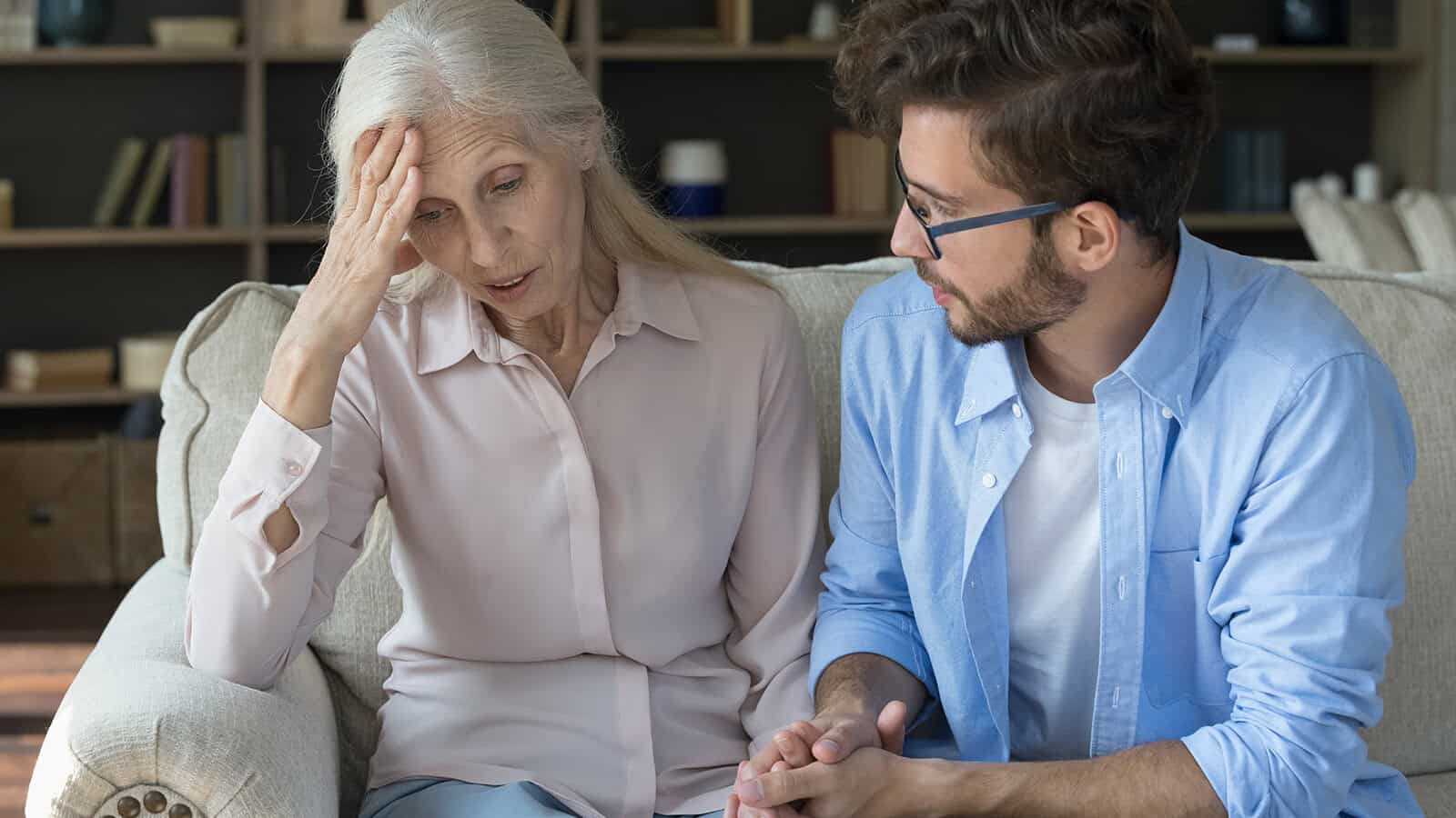 An older woman looks distressed, holding her head, while a younger man sits beside her, listening attentively and offering comfort in a living room.