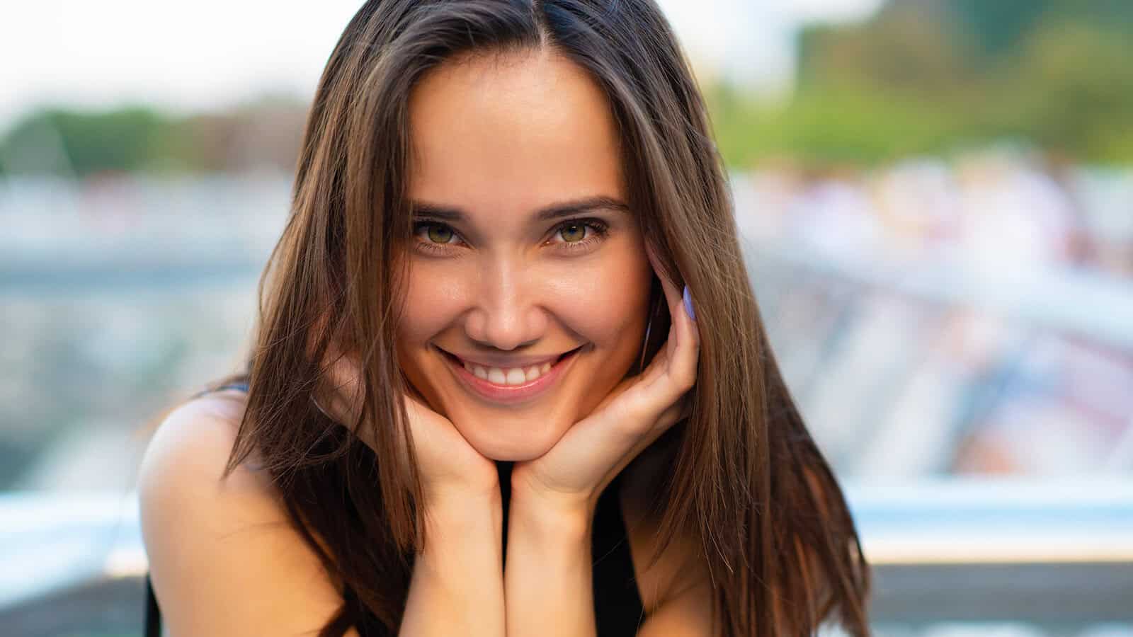 A woman with long brown hair smiles brightly, resting her chin on her hands, with a blurred outdoor background.