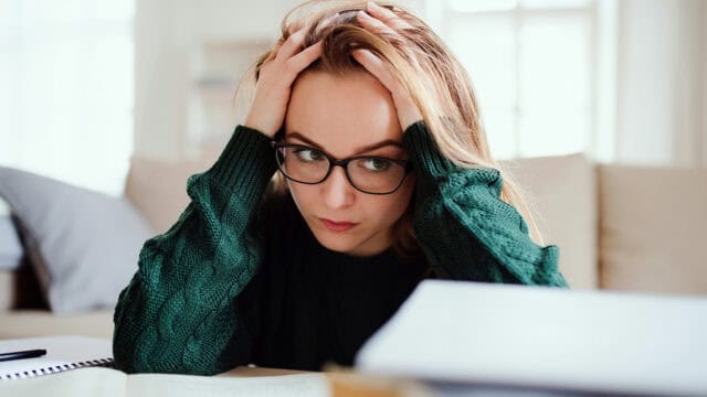 A woman wearing glasses and a green sweater sits at a desk, holding her head in her hands and looking stressed or worried while studying.