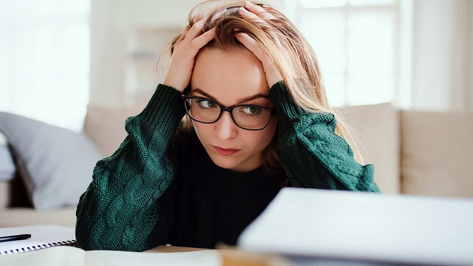 A woman wearing glasses and a green sweater sits at a desk, holding her head in her hands and looking stressed or worried while studying.