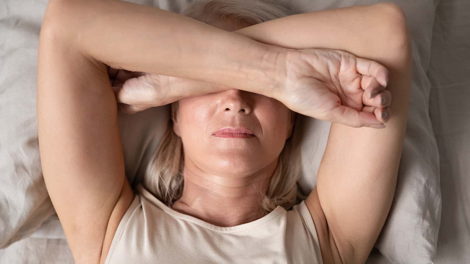A woman with light hair lies on her back in bed, covering her eyes with one arm and resting her other arm on her forehead, appearing tired or stressed.